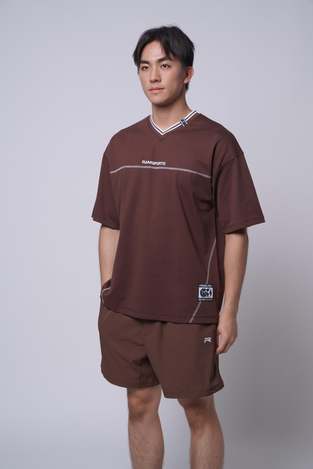 Asian male model standing with V-neck top with one hand on his pocket, wearing Ramsports MatchDay Core Short in brown