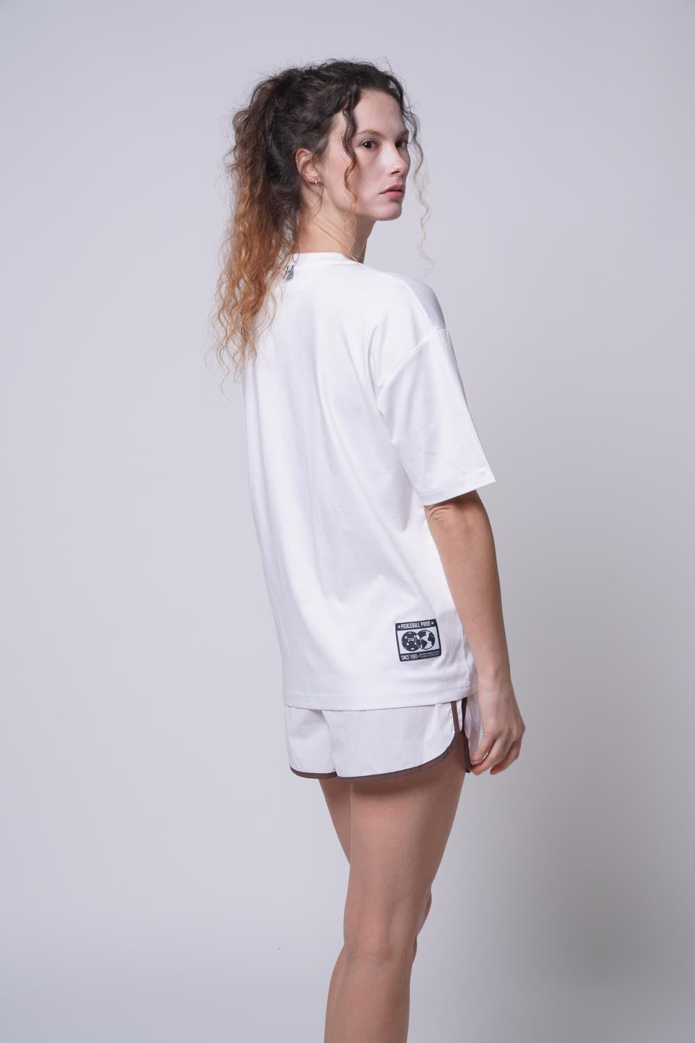 Female model facing away from the camera, looking to the side while wearing a white Ramsports Flow Panel Split-Hem Short and matching white shirt.