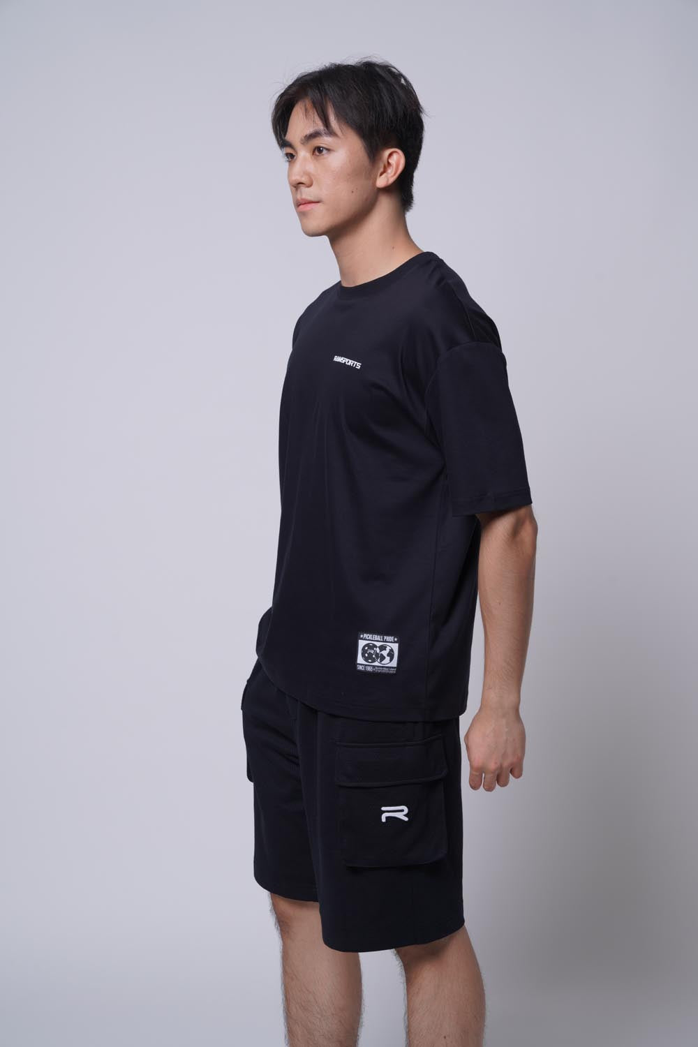 Male model wearing the black Ramsports Blueprint Back Graphic tee with black shorts against a gray background, body turned slightly to the side and looking away from the camera