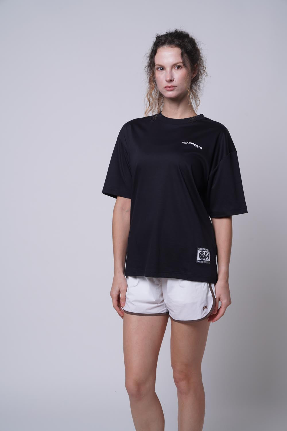 Female model wearing the black Ramsports Blueprint Back Graphic tee with white shorts against a gray background, hands at her sides, eyes looking sideways