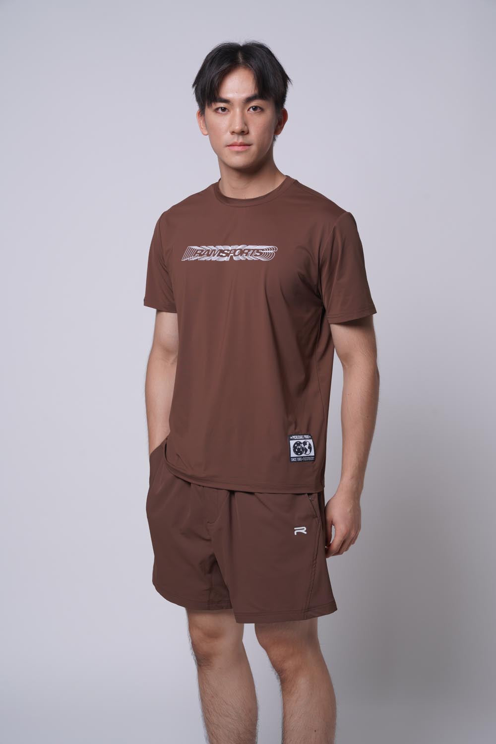 Male model wearing the brown Ramsports Playbook Court Diagram tee against a gray background with one hand in his pocket