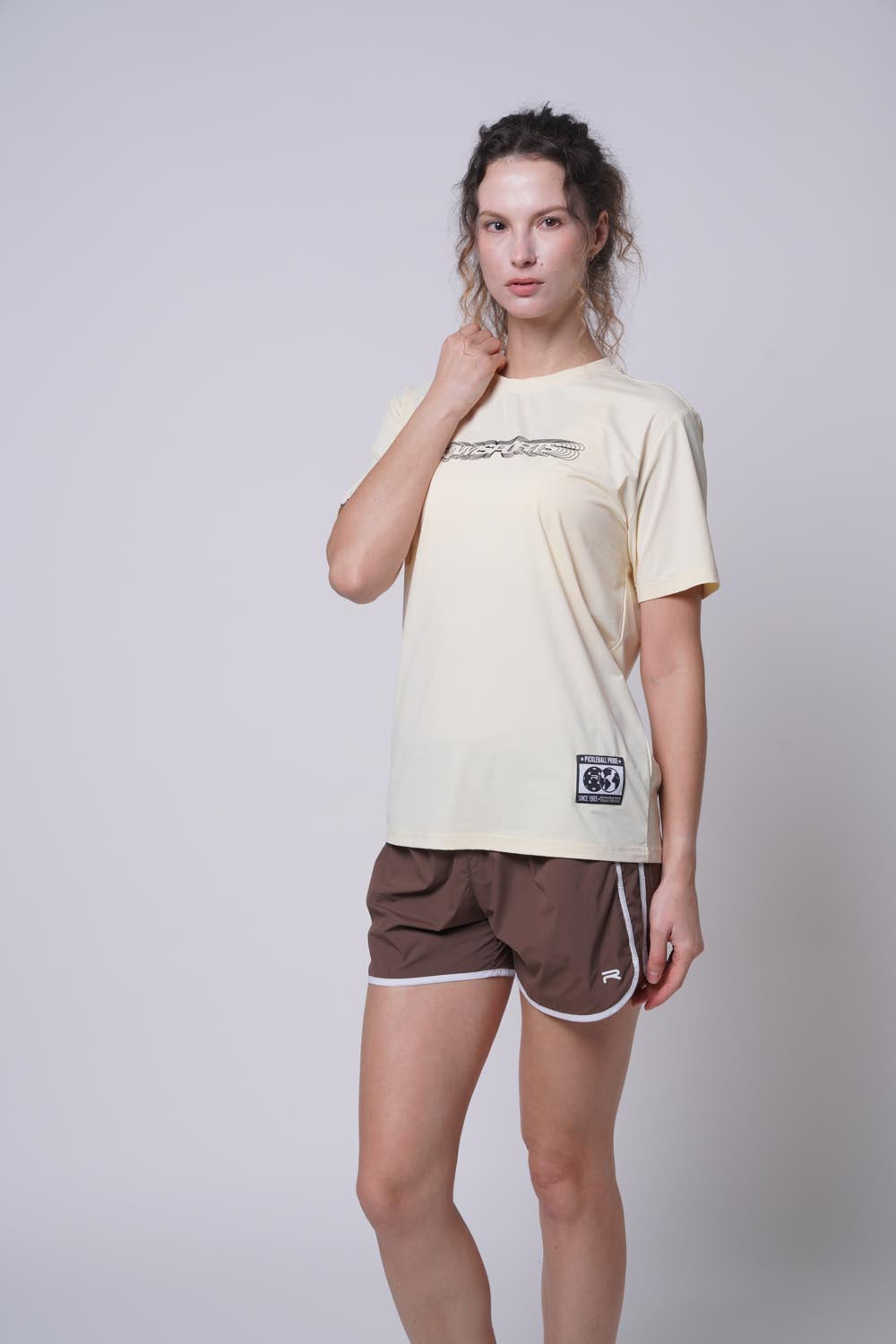 Female model wearing the beige Ramsports Playbook Court Diagram tee with brown shorts against a gray background, right hand resting on her neck