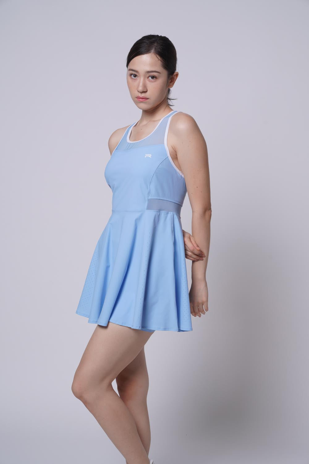 Asian female model with one arm behind her back and the other straight, one knee bent forward, wearing a blue Ramsports Racer Flare Dress.