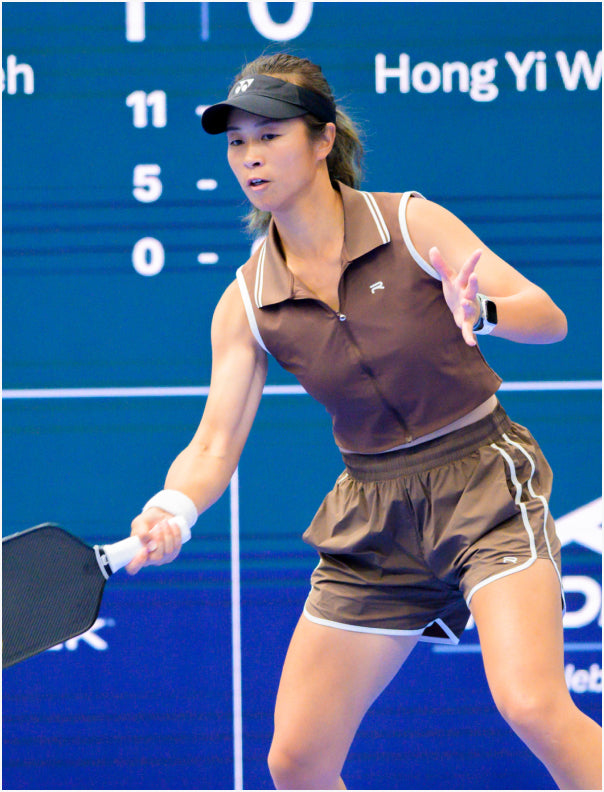 female pickleball player wearing the brown Ramsports Flow Panel Split-Hem Short as she's playing paddle on a blue court with branding in the background